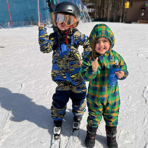 Two young children wearing colorful winter suits and skis, standing on a snowy slope, enjoying a day out in the winter sun.
