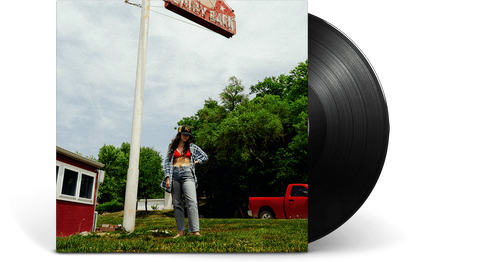 Person standing by an old sign with a classic record in the foreground, trees and a red truck in the background on a sunny day.