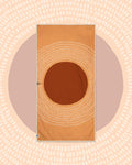 Alpenglow Wander & Travel Towel in orange with a large rust circle and dotted rays, on a peach and mauve sun print backdrop