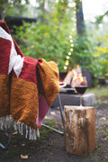Cozy Bernie Blanket draped over chair by campfire, glass of wine on log. Handwoven, made from recycled materials; perfect for relaxing. By Trek Light.