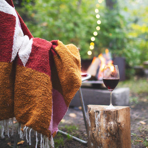 Cozy Bernie Blanket draped over chair by campfire, glass of wine on log. Handwoven, made from recycled materials; perfect for relaxing. By Trek Light.