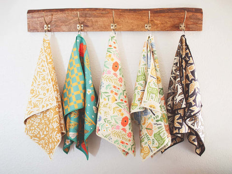 Colorful patterned quick-dry Trek Light Kitchen Dish & Tea Towels hanging on a wooden rack against a white wall.