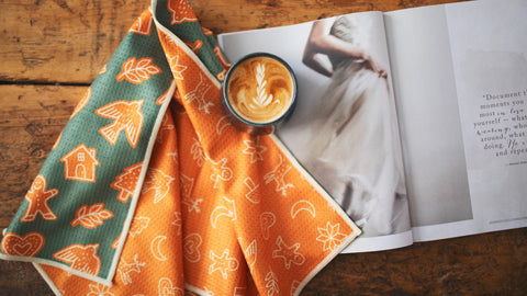 Latte art in a cup on a table with a colorful Trek Light kitchen towel and an open fashion magazine.