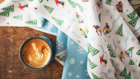 A cup of cappuccino on a wooden table next to a festive winter-themed Trek Light kitchen towel with skiers and snowflake patterns.