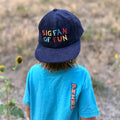 Child in blue shirt wearing a colorful 'Big Fan of Fun' Trek Light hat in a flat field, showcasing playful style for kids.