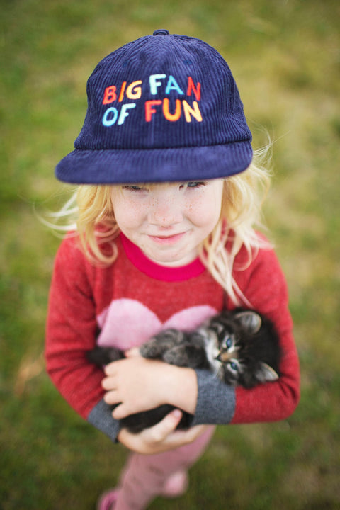 Child wearing a Big Fan Of Fun Kids Hat while holding a kitten in a park setting.