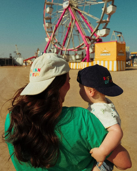 Adult and child wearing Big Fan Of Fun Hat - Twinsie Set at a fairground with a ferris wheel.