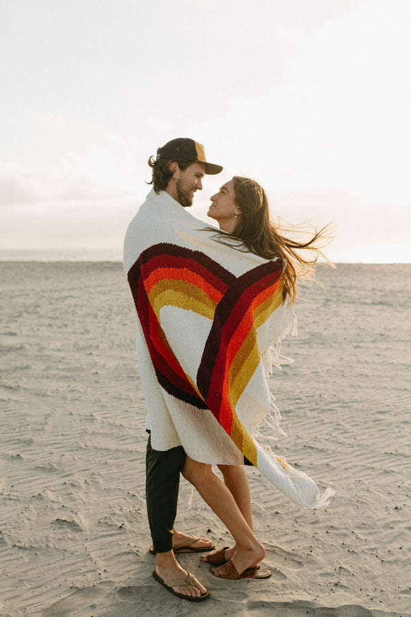 Couple wrapped in Trek Light's Bowie Blanket with vintage rainbow design, enjoying a beach moment.