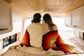 Couple wrapped in a Trek Light Bowie Blanket with rainbow design, enjoying a cozy moment in a wooden van interior.