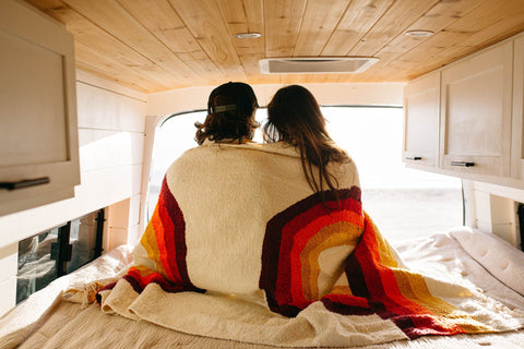 Couple wrapped in a Trek Light Bowie Blanket with rainbow design, enjoying a cozy moment in a wooden van interior.