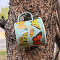 Butterfly Beauty Mug - Enamel 16oz Mug hanging on a tree, featuring beautiful butterfly designs.
