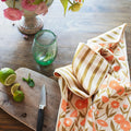 Calendula Flowers Kitchen & Tea Towel on a wooden counter with limes and a glass, showcasing vibrant floral design.
