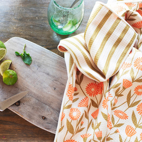 Calendula Flowers Kitchen & Tea Towel displayed on a wooden table with limes and a cocktail glass.