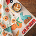 California Kitchen & Tea Towel with vibrant designs and eco-friendly fabric, shown with a latte on a wooden surface.