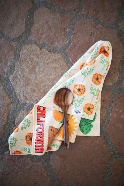 California Kitchen & Tea Towel with vibrant designs and a wooden spoon on a rustic stone surface.