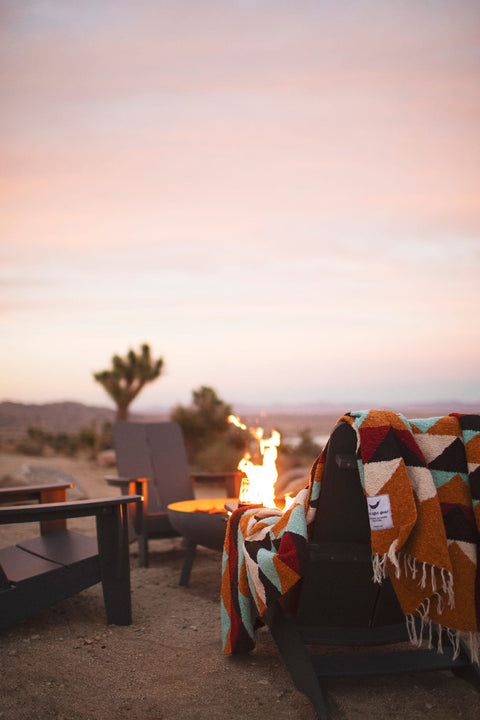 Beautiful handwoven Cascade Blanket made from recycled materials, draped over a cozy chair by a fire in a tranquil desert setting. By Trek Light.