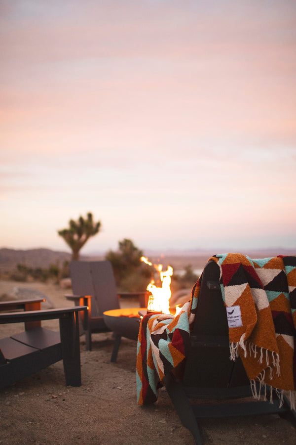 Beautiful handwoven Cascade Blanket made from recycled materials, draped over a cozy chair by a fire in a tranquil desert setting. By Trek Light.
