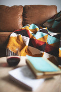 Beautiful handwoven Cascade Blanket made from recycled materials, featuring a colorful, cozy design on a sofa with a book and wine glass nearby. By Trek Light.