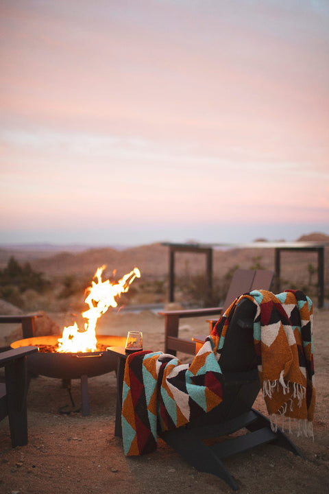 A colorful geometric patterned handwoven throw blanket is draped over a dark wooden chair next to a lit fire pit in an outdoor desert setting at sunset.
