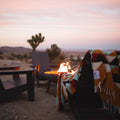 Beautiful handwoven Cascade Blanket made from recycled materials, draped over a cozy chair by a fire in a tranquil desert setting. By Trek Light.