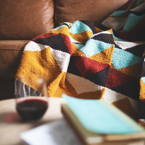 Beautiful handwoven Cascade Blanket made from recycled materials, featuring a colorful, cozy design on a sofa with a book and wine glass nearby. By Trek Light.