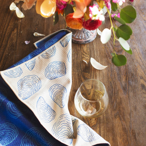 Coastal Oysters Kitchen & Tea Towel displayed with a floral arrangement and glass on a wooden table.