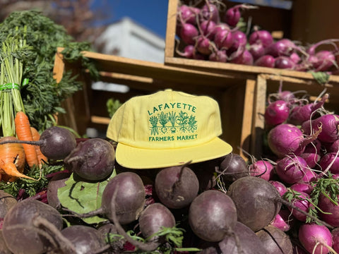 Yellow Lafayette Farmers Market hat surrounded by fresh vegetables including carrots, beets, and radishes.