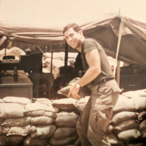 Man smiling while holding an object in front of a military tent and sandbags, wearing a watch and casual military attire.