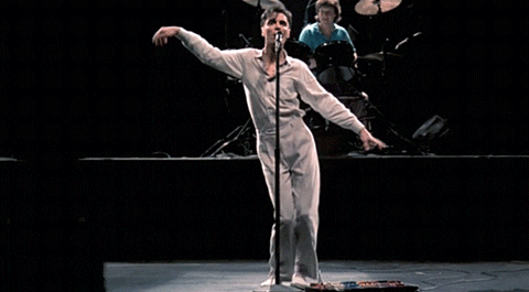A singer performing energetically on stage with a microphone, while a drummer plays in the background.