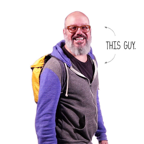 Smiling man wearing glasses and a grey sweater with purple sleeves, carrying a yellow backpack, labeled 'This Guy' by Trek Light.