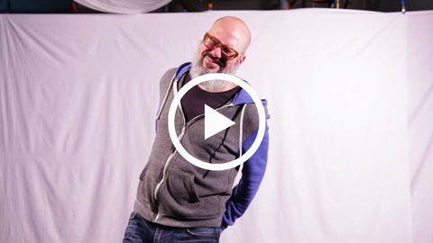 Person in hoodie smiling playfully against a white backdrop with video play button overlay.