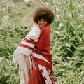 Person wrapped in a rust and white Divided Sky Blanket by Trek Light, handwoven with sustainable recycled materials in a lush field.