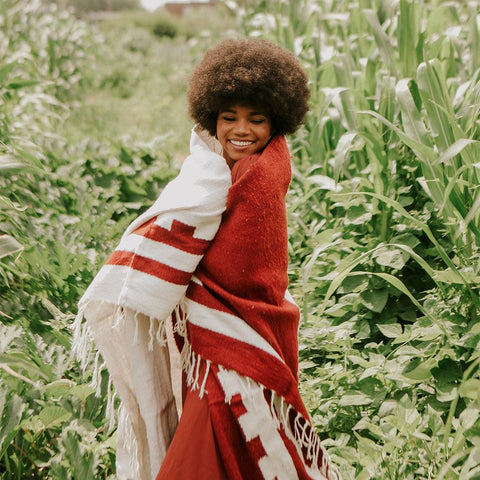 Person wrapped in a rust and white Divided Sky Blanket by Trek Light, handwoven with sustainable recycled materials in a lush field.