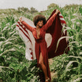 Woman joyfully holding a Trek Light blanket in a lush green field, wearing a flowing red dress.