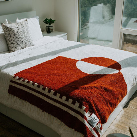 Rust and white divided sky blanket on bed, handwoven from recycled materials, by Trek Light. Eco-friendly and artisan-crafted.