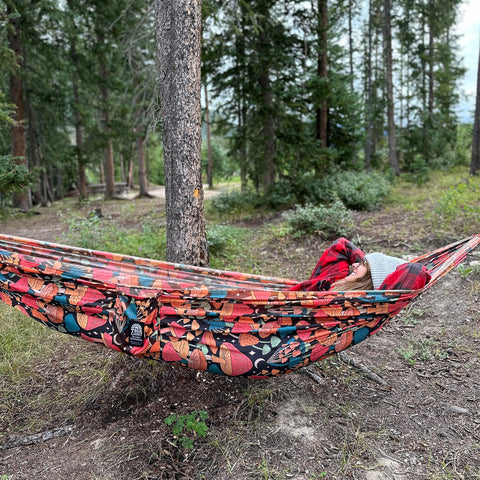 Person relaxing in a Trek Light double hammock with a midnight mushroom print, surrounded by trees in a serene outdoor setting.