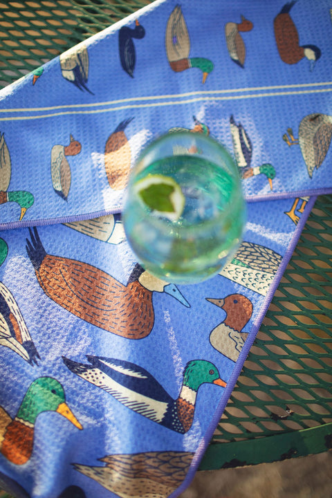 Ducks In A Row Kitchen & Tea Towel with colorful duck designs and a glass of drink on a table.
