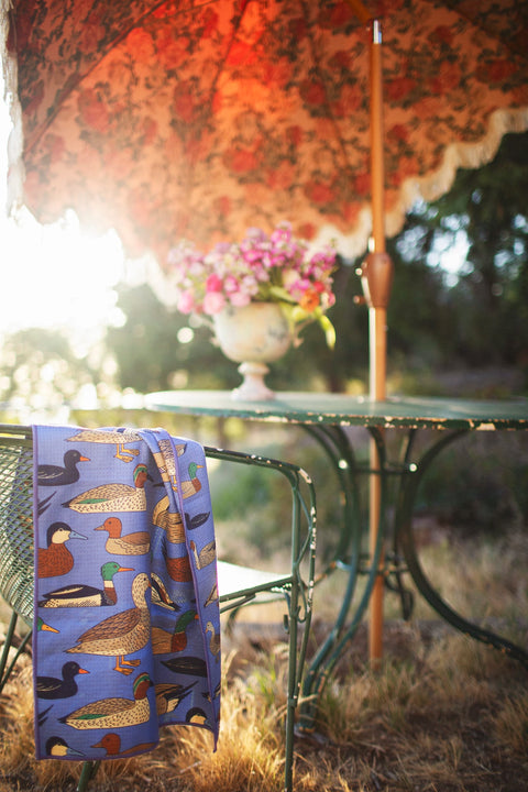 Ducks In A Row Kitchen & Tea Towel draped over a chair next to a table with flowers, showcasing vibrant duck designs.