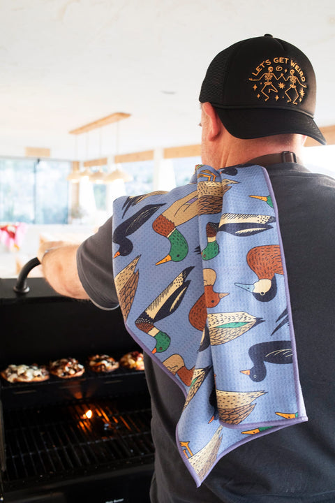 Person using Ducks In A Row Kitchen & Tea Towel while grilling, showcasing vibrant duck designs and practicality.