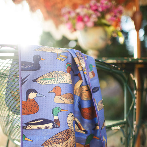 Ducks In A Row Kitchen & Tea Towel draped over a chair, featuring colorful duck designs against a blue background.