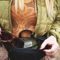 Woman opening Everyday Fanny Pack (Eclipse) showing spacious inner compartment with sunglasses, ideal for organized daily use.