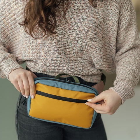 Person holding a yellow and blue travel pouch with a zipper, wearing a knit sweater and dark pants.