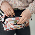 Person opening Everyday Fanny Pack in checkers design, showing multiple pockets for versatile use as hip or shoulder sling bag.