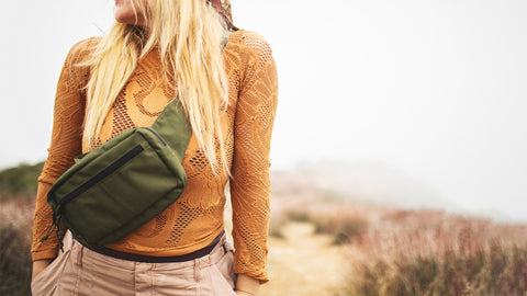 Woman wearing a chic orange top and a green fanny pack, walking along a misty trail in nature.