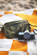 Everyday Fanny Pack (Fern) next to a Canon camera on a patterned blanket at the beach.