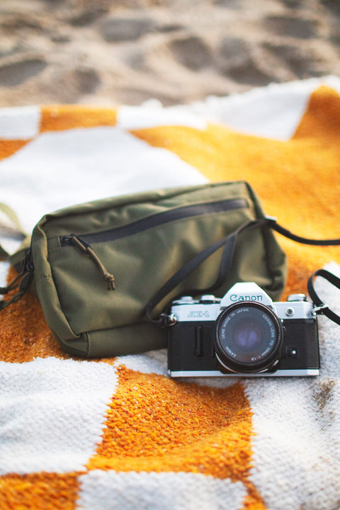 Everyday Fanny Pack (Fern) next to a Canon camera on a patterned blanket at the beach.