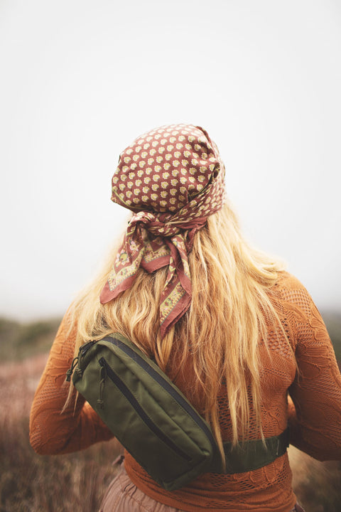 A woman wearing the Everyday Fanny Pack (Fern) in a lush outdoor setting, highlighted by her patterned scarf and hair.