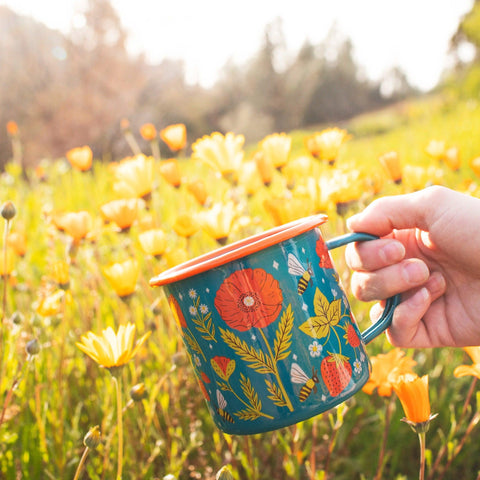 Garden Buzz Enamel Mug 16oz held in a field of yellow flowers, featuring vibrant floral design. Perfect for your next Trek Light adventure.