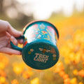 Colorful Trek Light Garden Buzz Enamel Mug 16oz in a field of orange flowers, perfect for outdoor adventures.