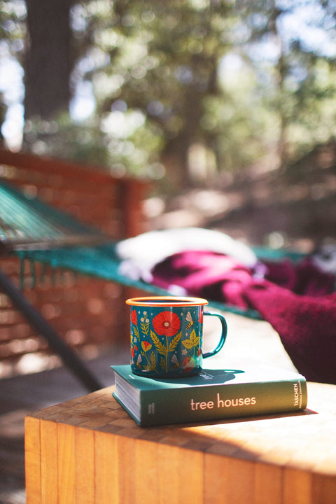 Garden Buzz Enamel Mug 16oz on a wooden table beside a book in a picturesque outdoor setting.